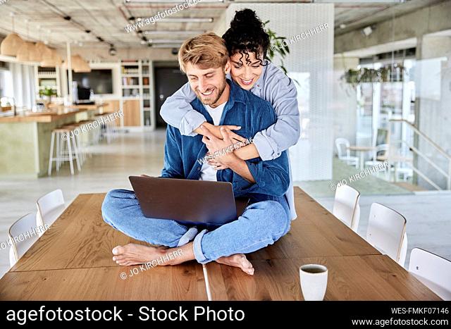 Smiling woman embracing boyfriend using laptop on table at loft