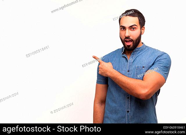 Excited bearded man in blue shirt pointing finger away at empty space, shocked surprised with advertisement area, recommending freespace