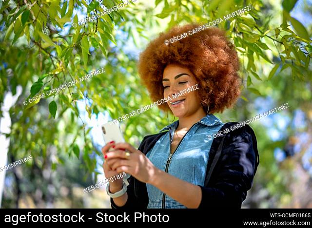 Smiling woman with frizzy hair using mobile phone at public park