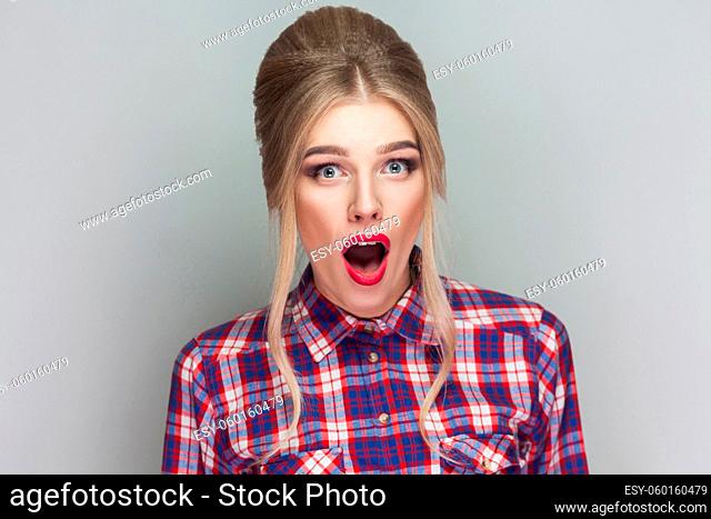 Amazed beautiful blonde girl with pink checkered shirt, collected updo hairstyle and makeup standing and looking at camera with shocked face and big eyes