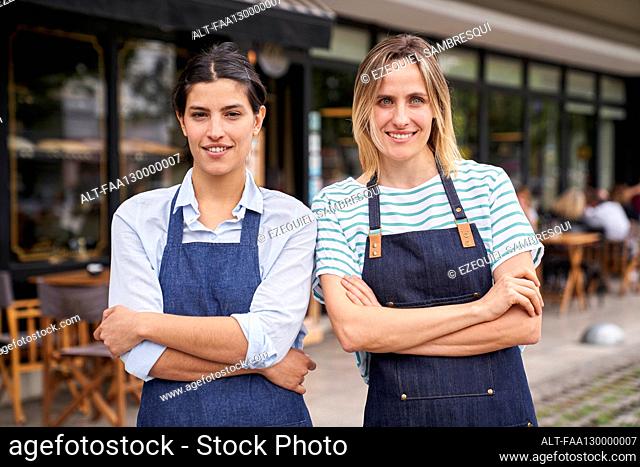Mid shot of two female restaurant entrepreneurs posing in front of their store