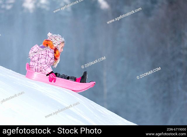 Kleines Mädchen in rosa Jacke und buntem Schild un drosa Schlitten rodelt eine Rodelbahn hinunter. Little girl sledging downhill an having fun