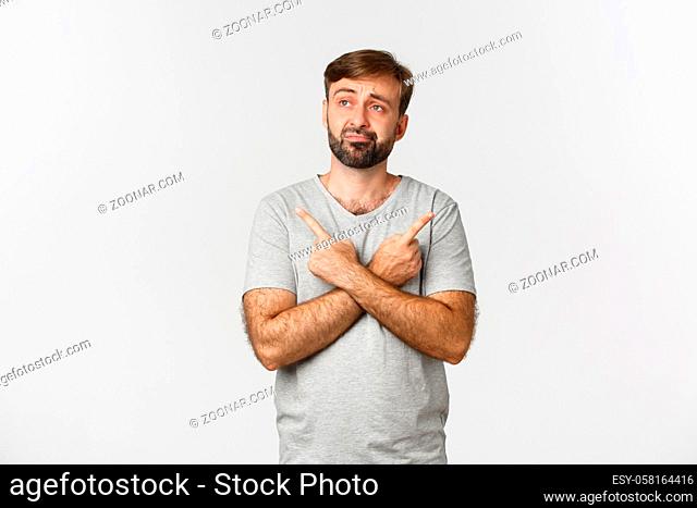 Portrait of indecisive beaded man in gray t-shirt, making decision, pointing fingers sideways at two choices, standing over white background