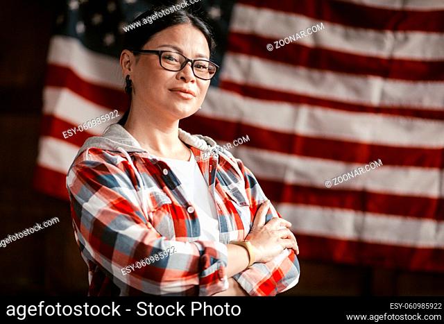 Asian Woman Smiles Patriotically On America's Flag Background, Beautiful Brunette Middle Aged In Glasses And Plaid Shirt Smiles With Crossed Arms On Her Chest
