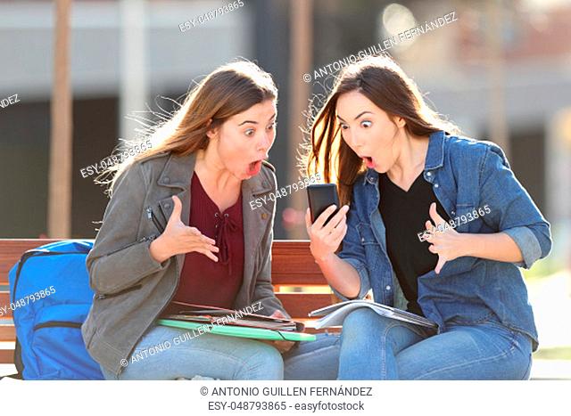 Two amazed students checking smart phone content sitting on a bench in a park