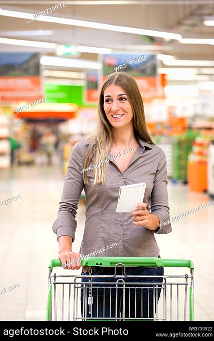 Smiling young woman shopping with trolley and checklist