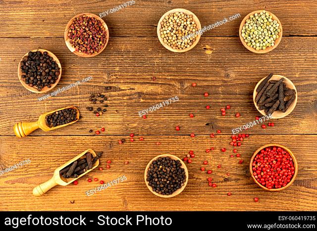 Seven small wooden bowls filled with different kinds of pepper and two wooden spice shovels and red peppercorns lie on a rustic wooden background with copy...
