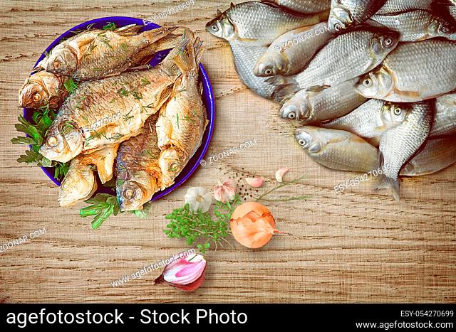 On the surface of the wooden table is a blue plate of fried fish, nearby is fresh, not cleaned fish and vegetables