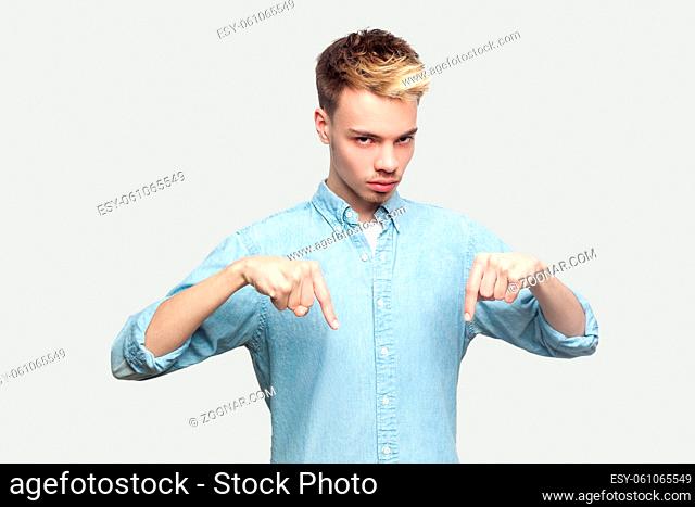 Here and right now. Portrait of serious handsome young man in light blue shirt standing, pointing his finger down, looking at camera with serious face