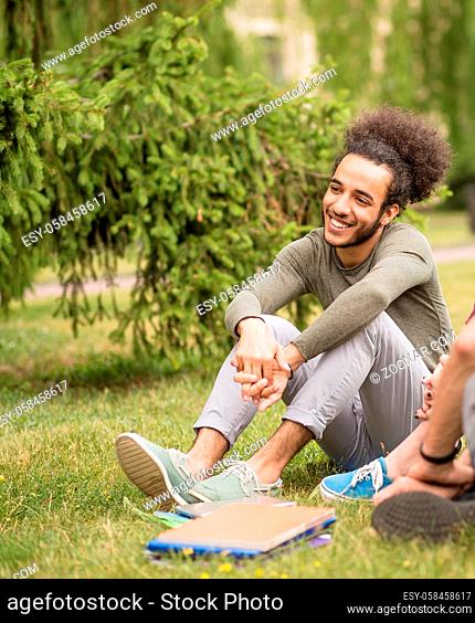 Young student man with stylish hair in campus. Boy smiling sitting at lawn in the University garden