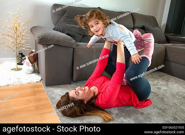 Mother stretched out on the ground plays with her daughter raising her up high and she smiles and raises her arms to fly