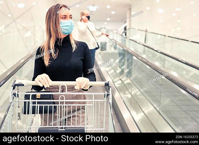Frau mit Mundschutz wegen Covid-19 mit Einkaufswagen auf einer Supermarkt Rolltreppe