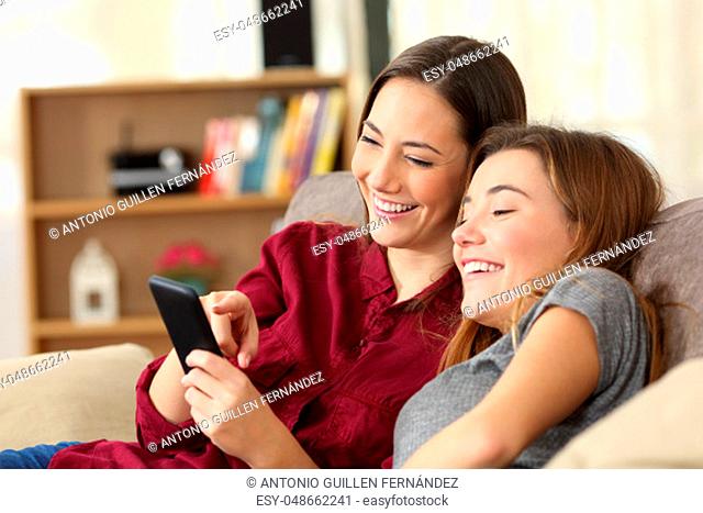 Two happy roommates checking smart phone content sitting on a couch in the living room at home