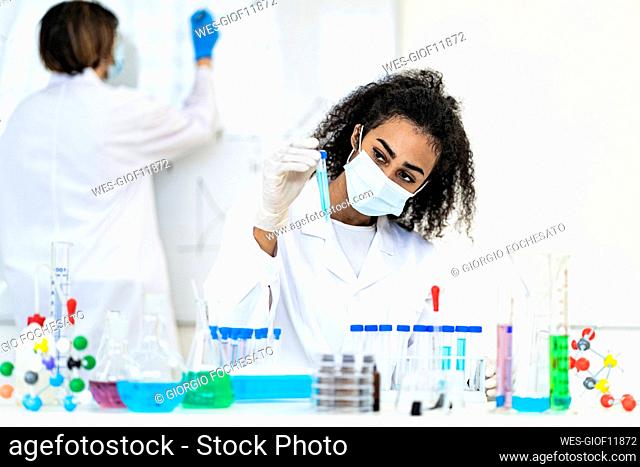 Female researcher examining liquid in test tube while coworker working in background at laboratory