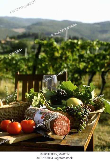 Table with salami, tomatoes, grapes in front of Tuscan vineyard