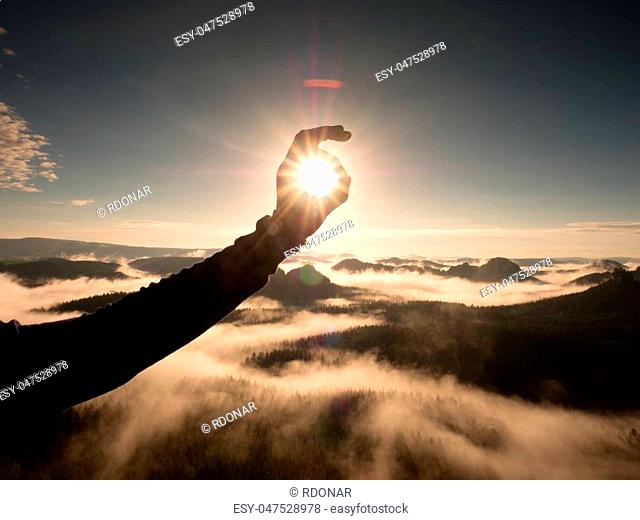 Man hand touch Sun. Misty daybreak in a beautiful hills. Peaks of hills are sticking out from foggy background, the fog is red and orange due to Sun rays