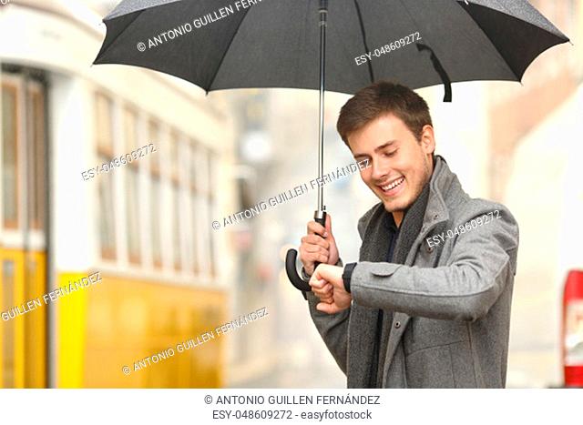Happy elegant man consulting information in a smartwatch waiting for public transportation in the street of an old town under an umbrella in a rainy day