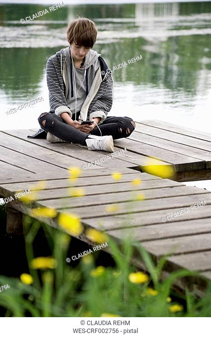 Teenage girl sitting on boardwalk at lake listening music with earphones
