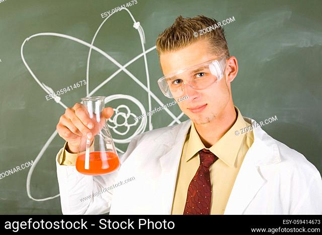 Young chemist in white apron. Holding beaker with orange liquid. Wearing goggles. Looking at camera, green background