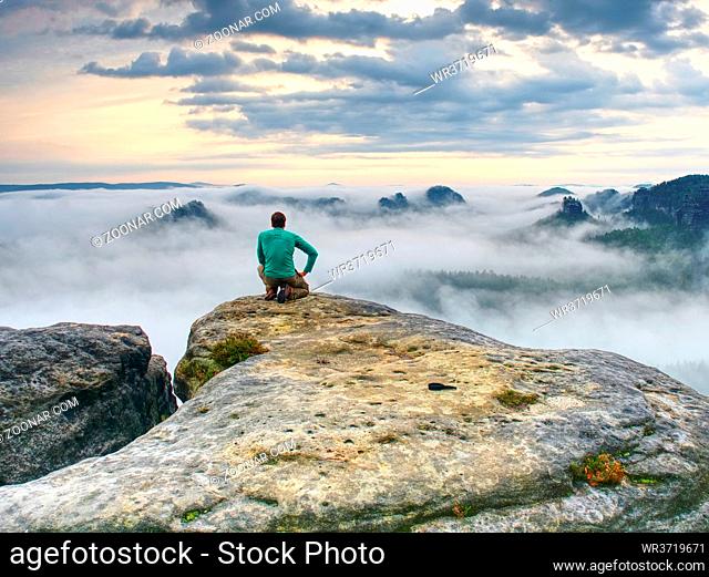 Professional photographer on knees at tripod looking into fall landscape and thinking. Hiker with tripod on cliff. Dreamy fall landscape with Sun at horizon
