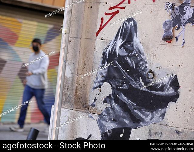22 January 2022, Spain, Palma: A man wearing a face shield walks past graffiti in Palma. The 14-day incidence in Mallorca is currently 3239 people tested...
