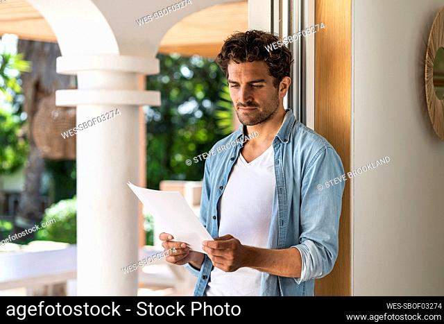 Serious man reading document while leaning on doorframe at home