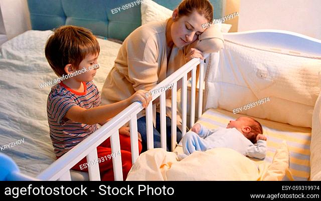 Older boy helping his tired mother rocking baby ccrib in bedroom at night. Children helping parents. Motherhood and sleepless nights
