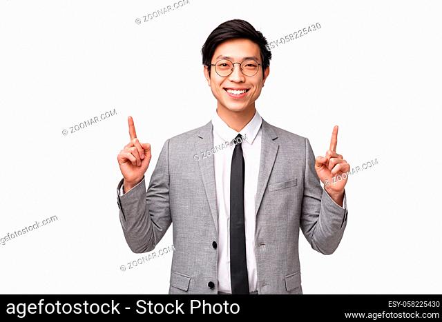 Waist-up portrait of successful smiling asian businessman in grey suit, pointing fingers up to show advertisement, presenting diagram or company chart