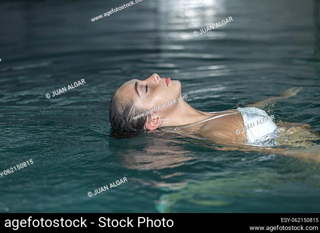 Relaxed young woman in white bra with wet hair closing eyes and floating in clean water