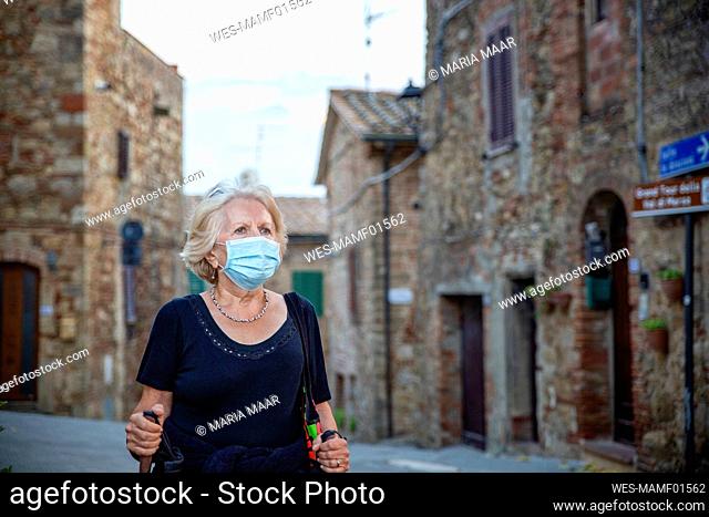 Senior woman wearing protective face mask holding hiking pole while standing in Italian city