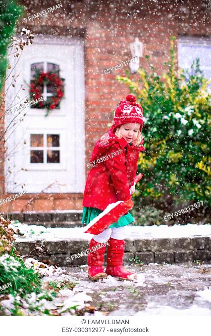 Little girl shoveling snow on home drive way. Beautiful house decorated for Christmas. Child with shovel playing outdoors in Xmas season