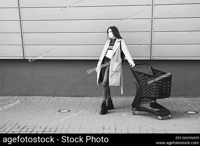 Young woman wearing protection face mask against coronavirus COVID-2019, novel coronavirus 2019-nCoV stands with shopping cart on supermarket department store...