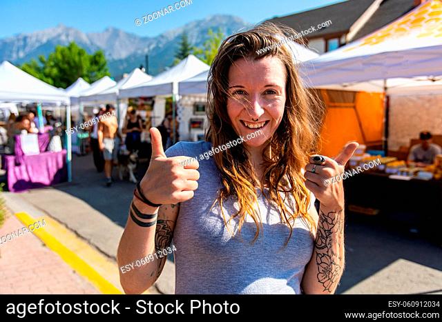 A healthy caucasian girl gives the thumbs up at local street fair for artisans and farmers, front portrait of happy shopper with market stalls behind