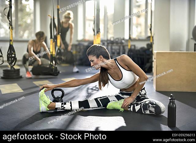 Pretty girl does stretch exercise on the floor in the gym. She wears black-white pants, white top and green sneakers. Woman holds her hands on the feet