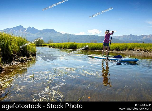 Caucasian woman paddle boarding at Rabbit Slough, with Pioneer Peak and Twin Peaks in the Chugach Mountains in the background on a sunny