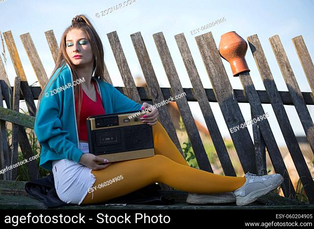 A beautiful country girl in bright clothes sits on a wooden bench with a cassette recorder. Woman in the style of the 90s