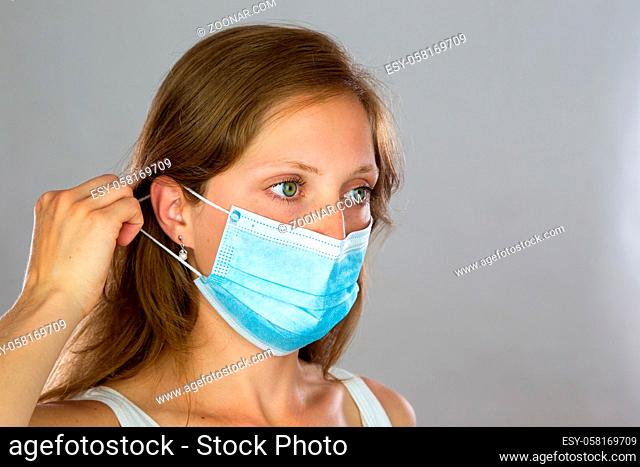 Female model in white shirt attaching surgical face mask on light background. Concept of prevention and protection in medicine