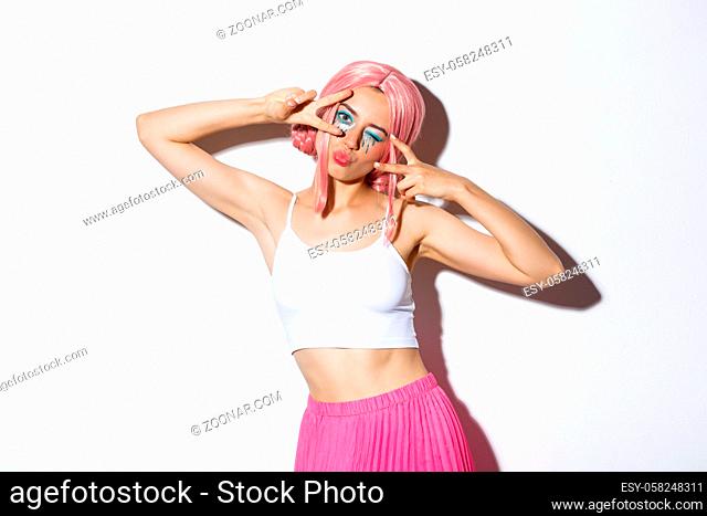 Carefree stylish girl with pink wig and bright makeup, having fun on party, celebrating and dancing, showing peace signs, standing over white background