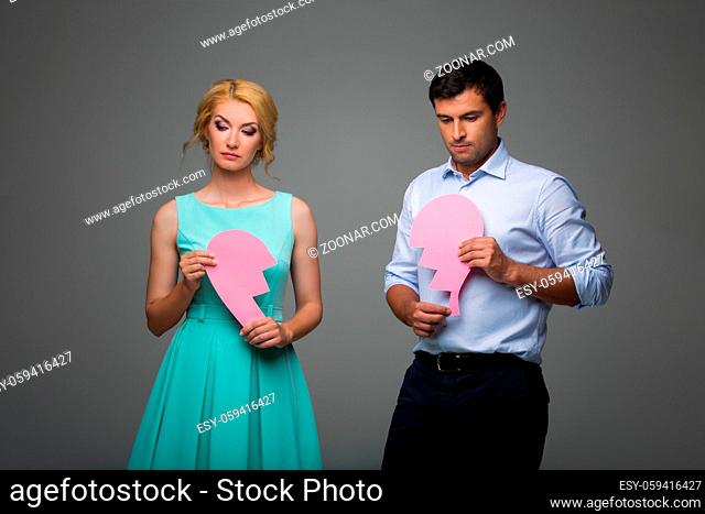 Beautiful young couple holding big pink broken heart. Blond woman in mint dress and handsome man in shirt and trousers standing on grey background
