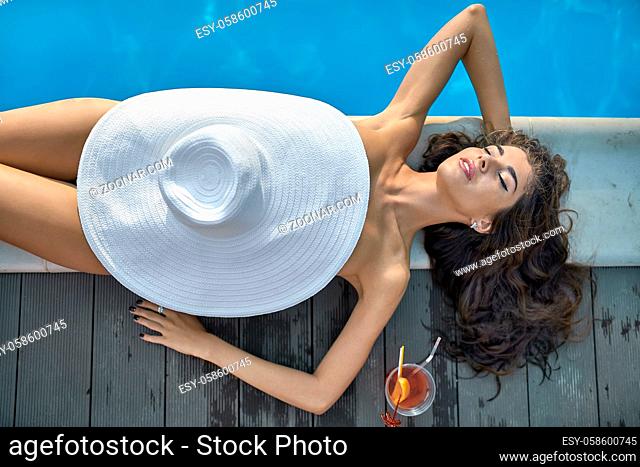 Sensual girl with closed eyes and parted lips lies with a big hat on the pool's edge outdoors. Next to her there is an orange cocktail