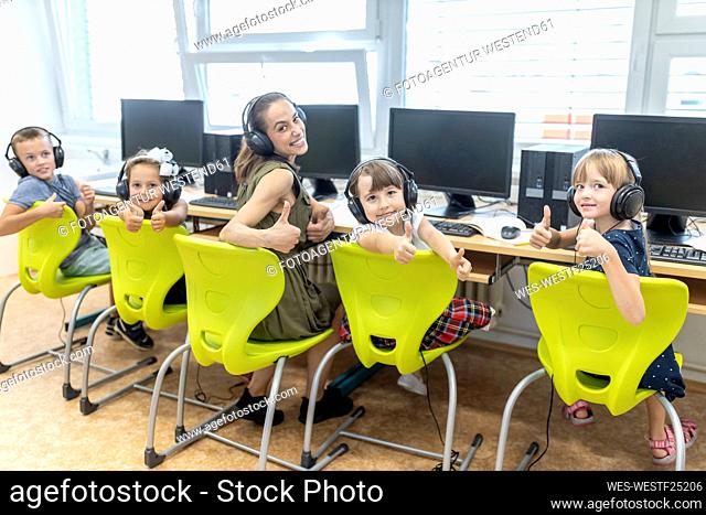 Teacher and students showing thumbs up sitting on chair in computer class at school