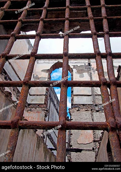 view through rusty iron bars on a window of a ruined collapsing building with crumbling walls and open to the sky