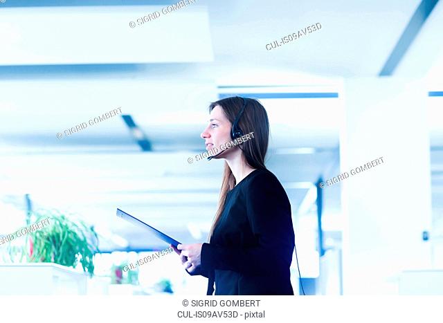 Side view of young woman in office wearing telephone headset, holding digital tablet looking away