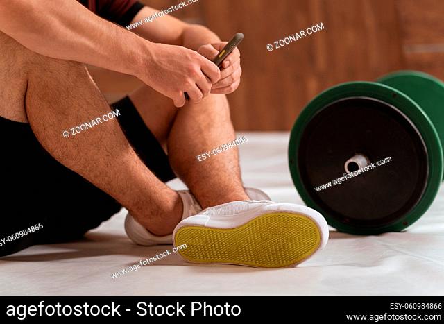 Close up. Young man having a break sitting on a floor with black and green tone fitness barbell, equipment for weight training concept