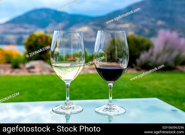 pair of wine glasses filled with red and white wines, selective focus and close up view against yard on Okanagan Lake and mountains background