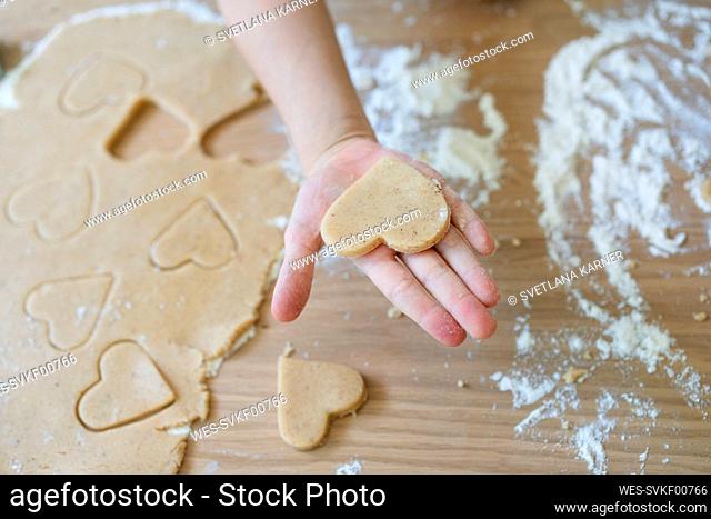 Hand of girl holding heart shaped cookie dough on table at home