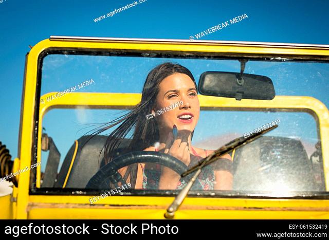 Happy caucasian woman sitting in beach buggy by the sea putting on lipstick