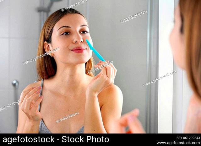 Beautiful young caucasian woman shaving her face by razor at home. Pretty woman using razor on bathroom