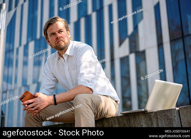 Businessman with laptop and smartphone having a break in the city sitting on a wall