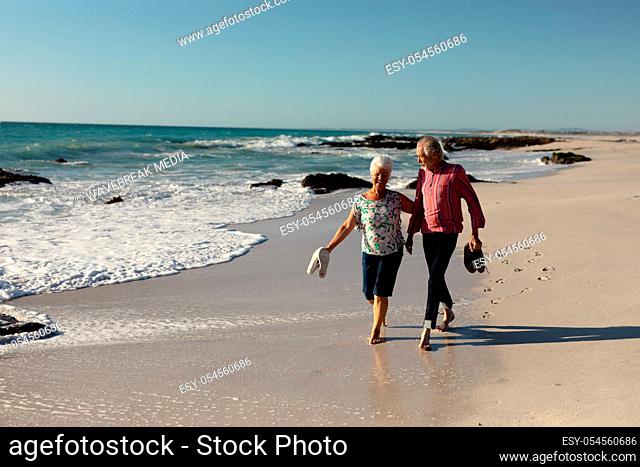 Front view of a senior Caucasian couple at the beach in the sun, holding their shoes and walking barefoot, smiling and talking